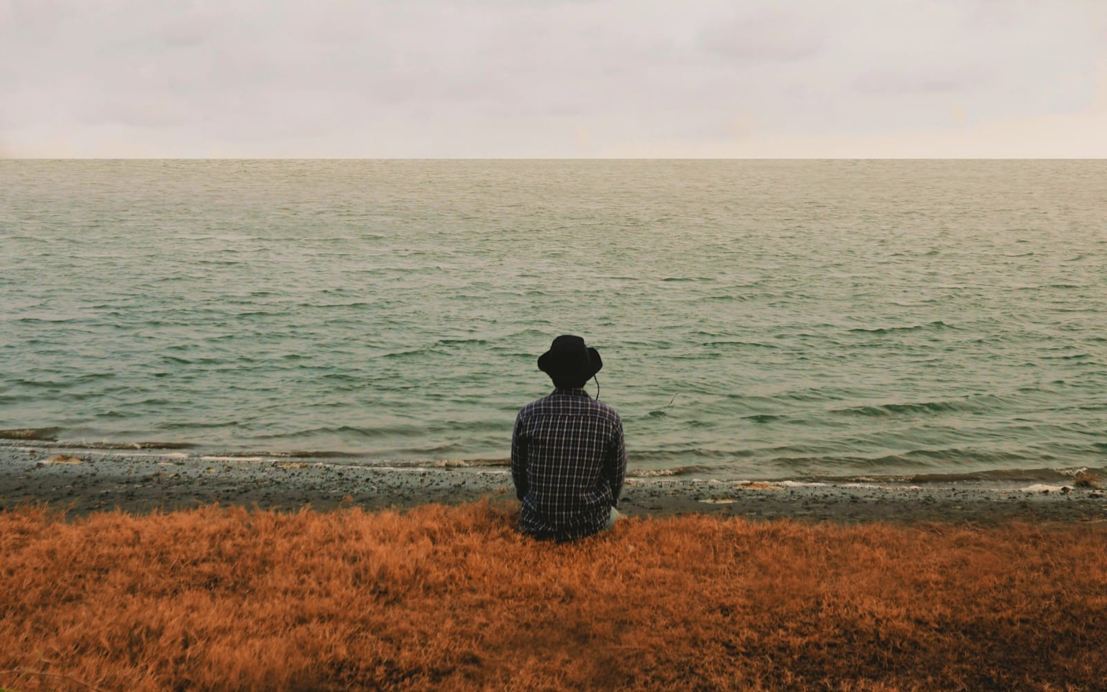 A man in a plaid shirt and bucket hat sits alone on a grassy shore, facing a vast grey-green lake under a cloudy sky.